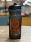 A dark gray water bottle with an orange and white 'Spoke Cycles CC' logo, sitting on a wooden table with a blurred cafe background.